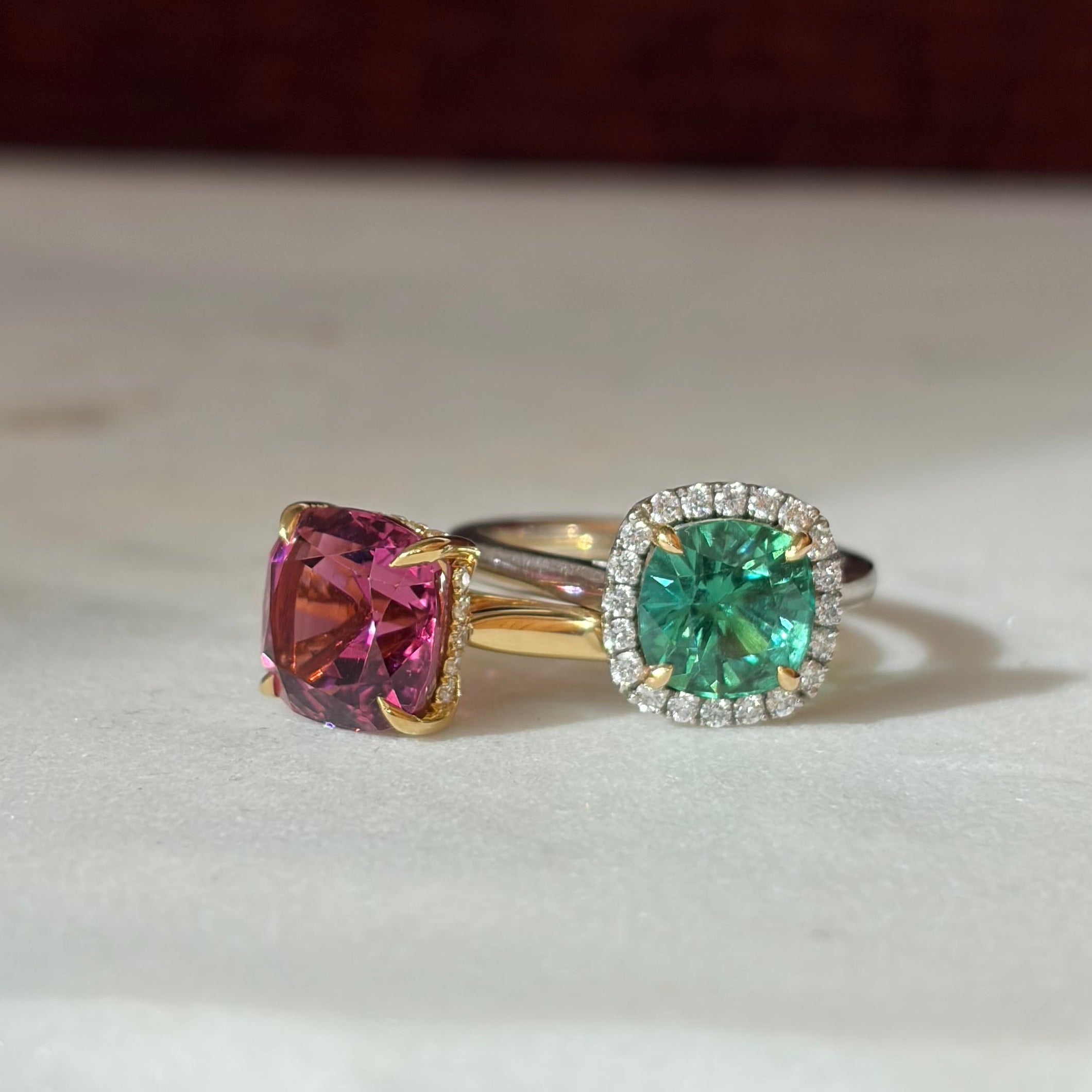 Two rings set on white marble. Green tourmaline with diamond halo se in platinum. Pink tourmaline solitaire with hidden diamond halo.