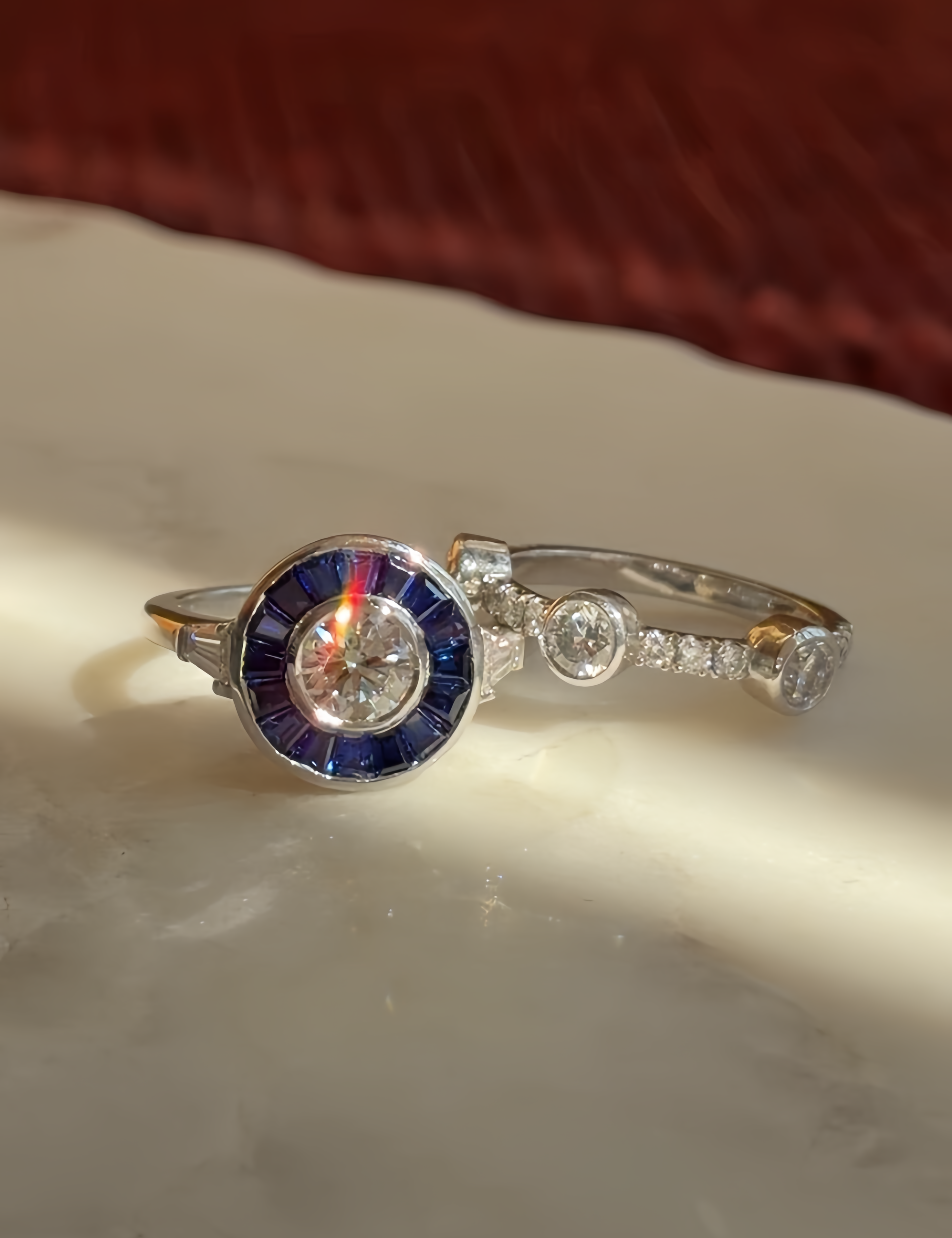 Vintage Inspired sapphire target ring, diamond centre with blue sapphire surround. Trapeze shoulders, set in platinum. Beside lies a 3 stone round brilliant diamonds, bezel set and in platinum on a diamond band.